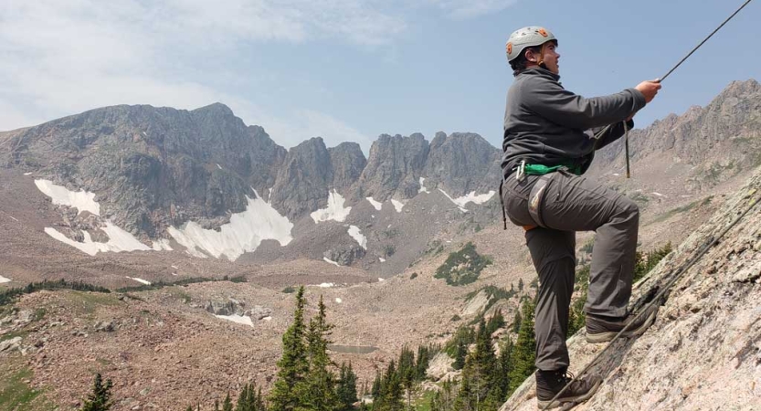 a person wearing safety gear holds onto a rope on a steep incline. Behind them are rocky mountains. 